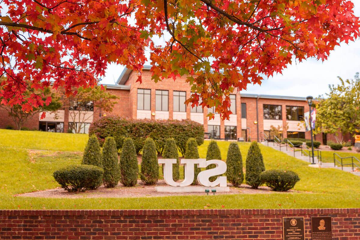 Shenandoah University quad in fall with red maple in the foreground and SU initial statue in center of the photo, in front of Wilkins Administration Building.